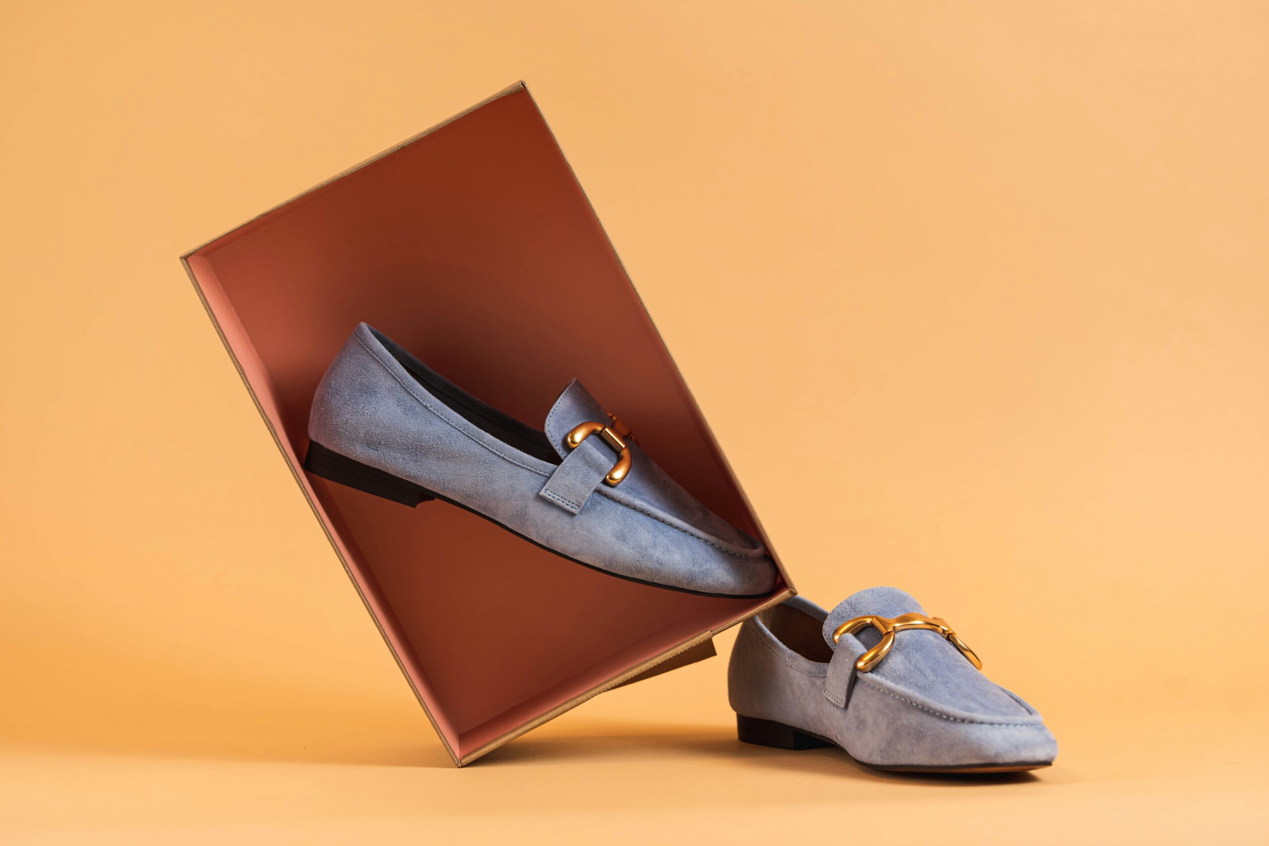 Elegant suede loafers with gold horsebit detailing styled in a modern geometric product display against a warm neutral background.
